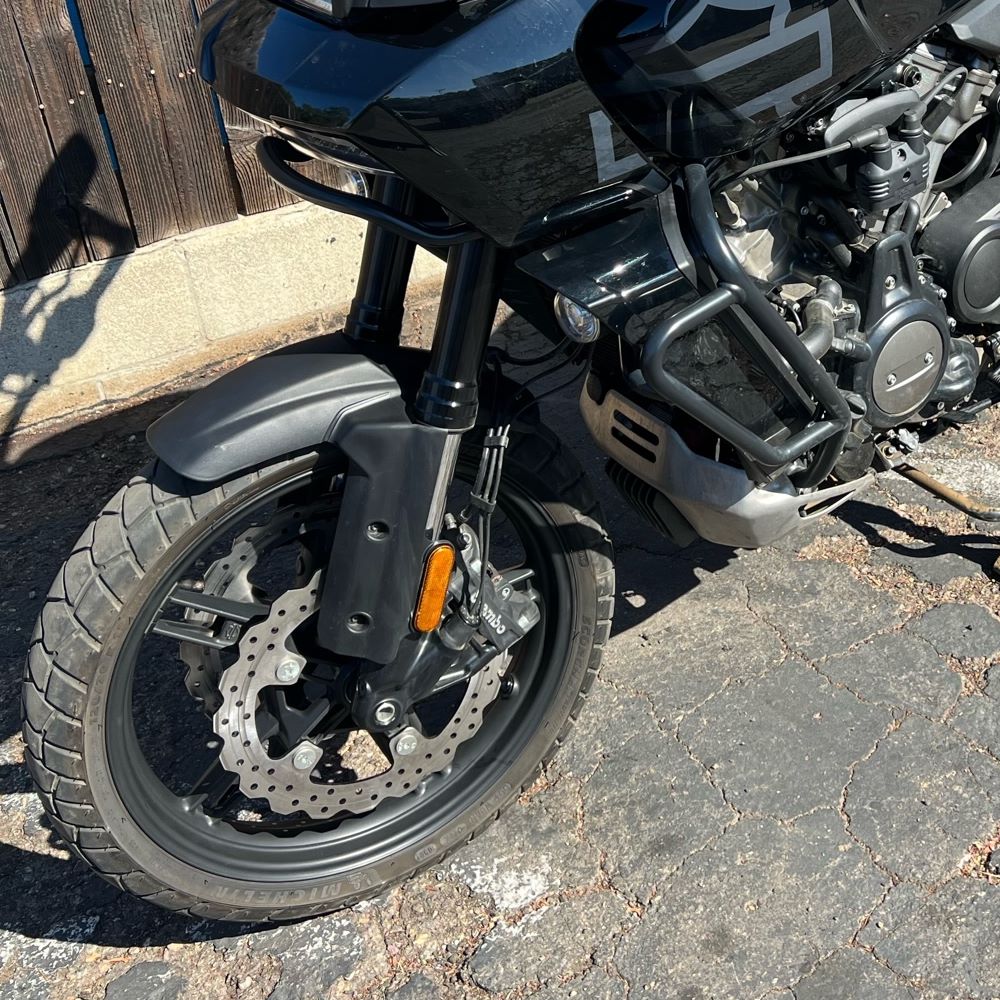 A black motorcycle, the TC Bros. Profile™ Front Floating Brake rotor for 2021-up Harley Pan America Mag Wheels, is parked in front of a fence.