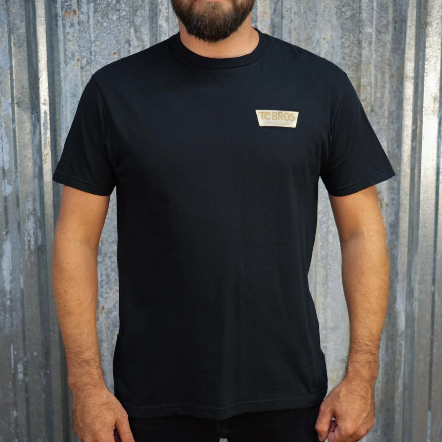 A person wearing the Genuine Quality - Black Tee by TC Bros., crafted from soft and comfy ring-spun cotton, featuring a small logo on the front, stands against a corrugated metal background.