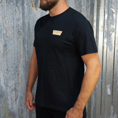 A person wearing the Genuine Quality - Black Tee by TC Bros., crafted from soft and comfy ring-spun cotton, featuring a small logo on the front, stands against a corrugated metal background.