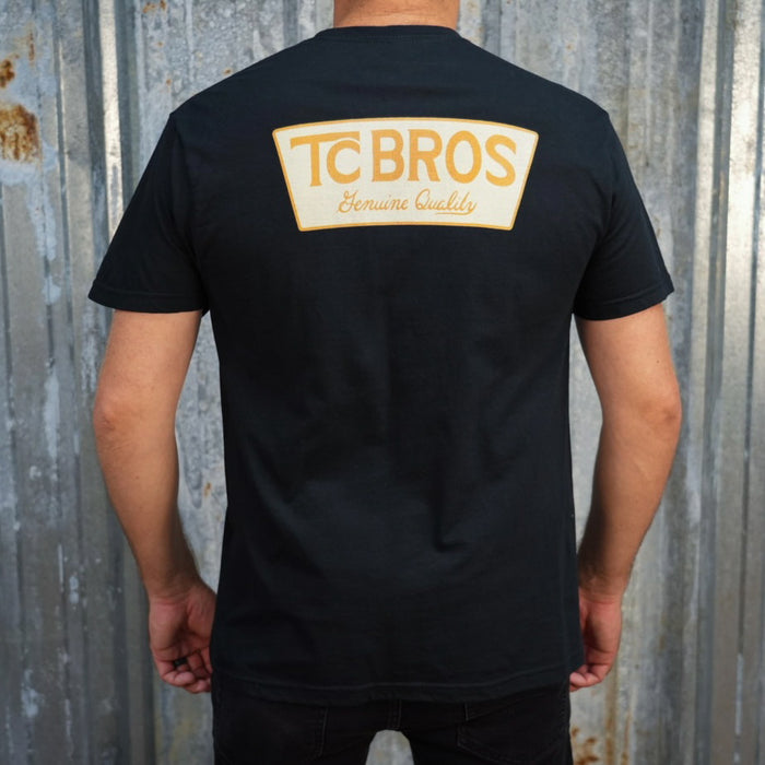 A person wearing the Genuine Quality - Black Tee by TC Bros., crafted from soft and comfy ring-spun cotton, featuring a small logo on the front, stands against a corrugated metal background.