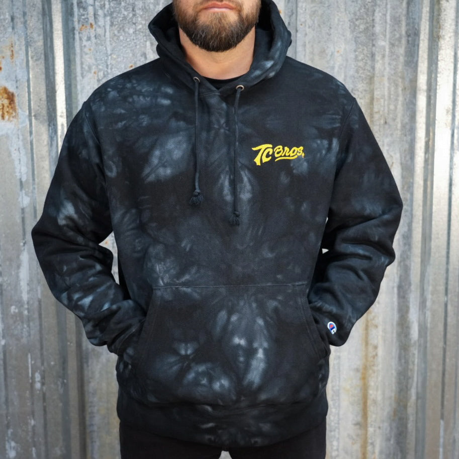 A black and gray camouflage patterned TC Bros. tie-dye hoodie with a yellow logo on the left chest.