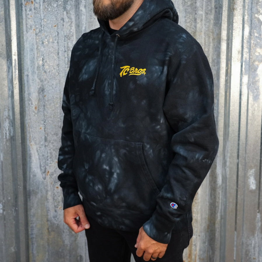 A black and gray camouflage patterned TC Bros. tie-dye hoodie with a yellow logo on the left chest.