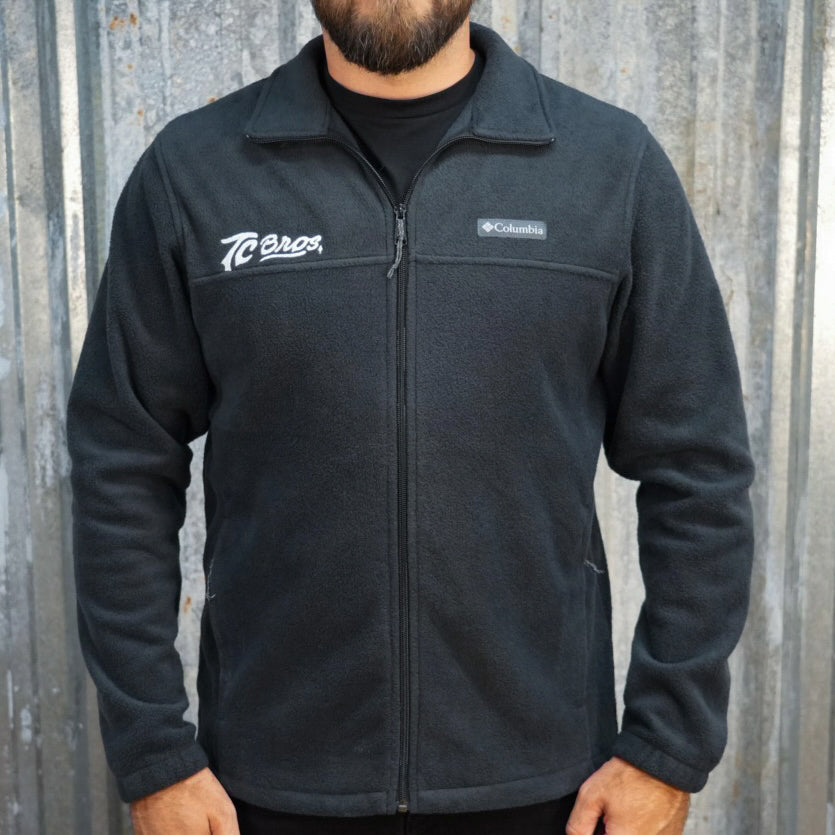 TC Bros. fleece zip jacket - Black with a high collar and zippered front, featuring white embroidered logos on the left chest.