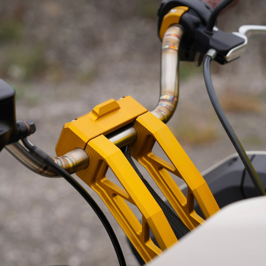 Close-up of a motorcycle's handlebars featuring yellow brackets and TC Bros. Pro Series Pullback Truss Riser for Harley M8 Softail Digital Gauge, highlighting American craftsmanship in the detailed control buttons and grips, with rear-view mirrors reflecting the paved surface below.