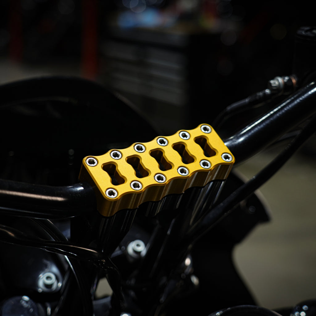 Close-up of the TC Bros. Top Clamp for Pro Series Modular Riser by TC Bros., gold anodized billet aluminum, mounted with multiple bolts on black Harley-Davidson handlebars in a workshop.