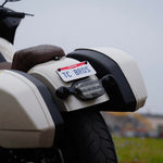 The TC Bros. Laydown License Plate Mount for Harley-Davidson features a black metal fender bracket with four screw holes and includes screws, washers, lock washers, nuts, and rubber grommets—perfect for secure mounting.