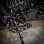 Close-up of TC Bros. Pro Series MX Slim Floorboards for 1984-Up Harley-Davidson Touring, featuring CNC-machined billet aluminum and visible bolts, mounted on a black Harley-Davidson motorcycle on a concrete floor.