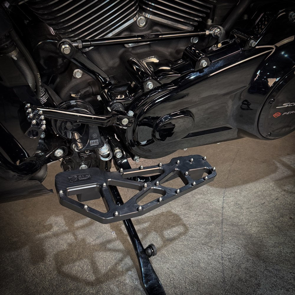 Close-up of TC Bros. Pro Series MX Slim Floorboards for 1984-Up Harley-Davidson Touring, featuring CNC-machined billet aluminum and visible bolts, mounted on a black Harley-Davidson motorcycle on a concrete floor.