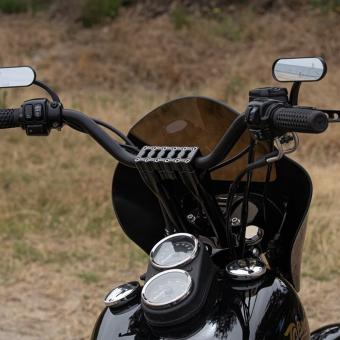 Close-up of a motorcycle’s handlebars equipped with TC Bros. Pro Series Modular Risers for Harley-Davidson, billet mirrors, and dashboard gauges; dry grass and shrubs are visible in the background.