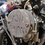 Close-up of a mechanical component featuring a "Pangor Cycles" branding on a circular metal plate. Various bolts, valves, and metal parts are visible, suggesting a part of an engine assembly, possibly connected to the Cheap Sucker Bird Catcher/Air Cleaner for S&S Super E Carburetors.