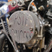 Close-up of a mechanical component featuring a "Pangor Cycles" branding on a circular metal plate. Various bolts, valves, and metal parts are visible, suggesting a part of an engine assembly, possibly connected to the Cheap Sucker Bird Catcher/Air Cleaner for S&S Super E Carburetors.