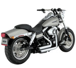A silver and black cruiser-style motorcycle with chunky tires, a chrome Vance & Hines Short Shots Exhaust System for 06-09 Fat Bob, Dyna, Low, Wide & Super Glide, and a single seat, viewed from the rear left side.
