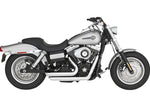 A silver and black cruiser-style motorcycle with chunky tires, a chrome Vance & Hines Short Shots Exhaust System for 06-09 Fat Bob, Dyna, Low, Wide & Super Glide, and a single seat, viewed from the rear left side.