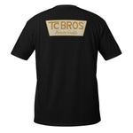 A person wearing the Genuine Quality - Black Tee by TC Bros., crafted from soft and comfy ring-spun cotton, featuring a small logo on the front, stands against a corrugated metal background.