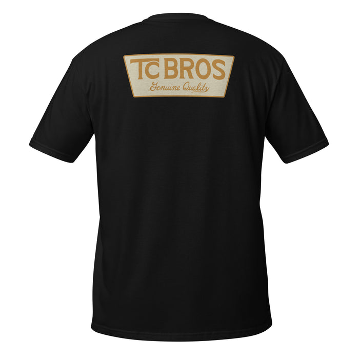 A person wearing the Genuine Quality - Black Tee by TC Bros., crafted from soft and comfy ring-spun cotton, featuring a small logo on the front, stands against a corrugated metal background.