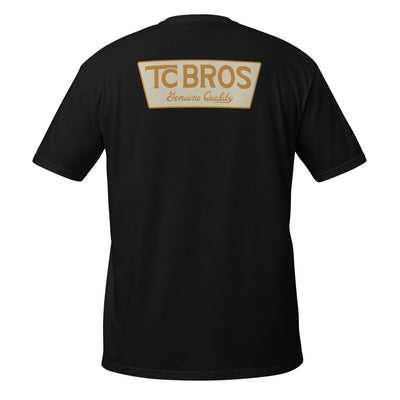 A person wearing the Genuine Quality - Black Tee by TC Bros., crafted from soft and comfy ring-spun cotton, featuring a small logo on the front, stands against a corrugated metal background.