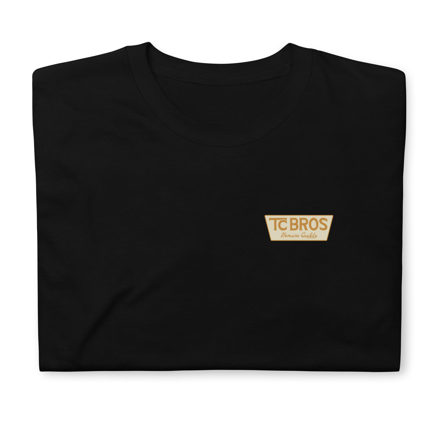 A person wearing the Genuine Quality - Black Tee by TC Bros., crafted from soft and comfy ring-spun cotton, featuring a small logo on the front, stands against a corrugated metal background.