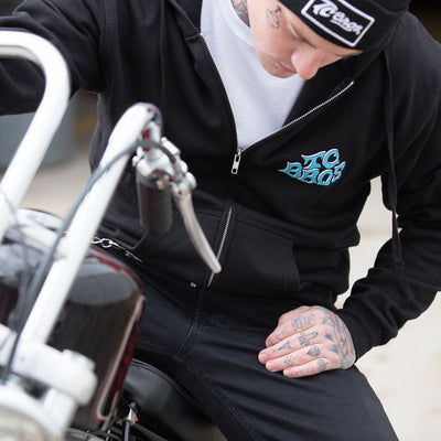 A man sitting on a motorcycle wearing a black TC Bros. Eagle Zip Hoodie featuring the TC Bros. Eagle design.