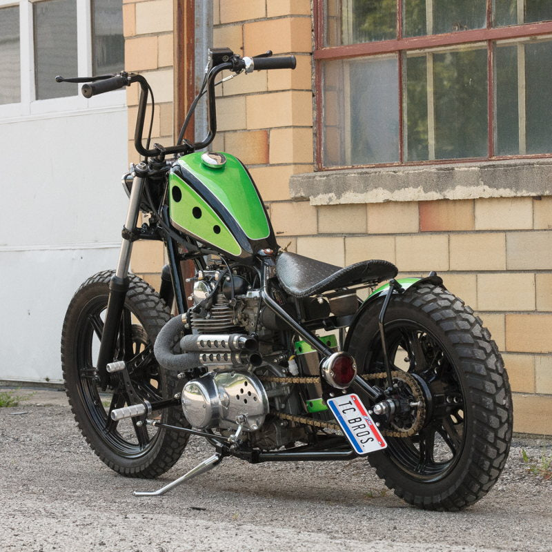 A green and black motorcycle with a Moto Iron® 2.5 Gal. Frisco Mount Sportster Bobber Gas Tank parked in front of a building.