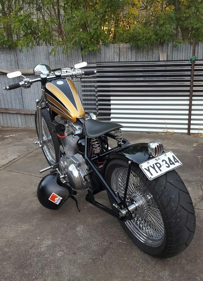 A black and gold TC Bros. Harley Sportster Hardtail kit motorcycle parked in front of a fence, showcasing wide wheel and tire applications.