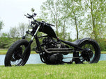 A motorcycle parked in the grass near a lake with a black custom Yamaha XS650 "Ya Mama" Exhaust System By Pandemonium (Fits 1974-1982 XS650).