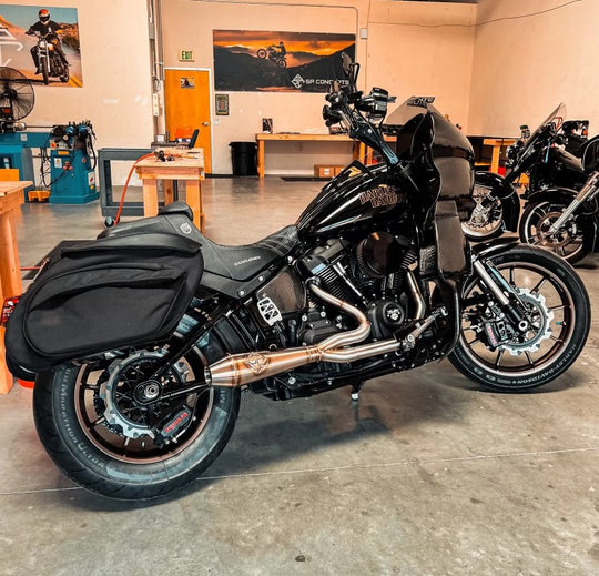 The SP Concepts Big Bore Exhaust for M8 Softail ST & Sport Glide (2018-Present) in stainless steel, featuring welded joints and a hexagonal logo emblem, rests on a metallic work surface, highlighting its premium finish.