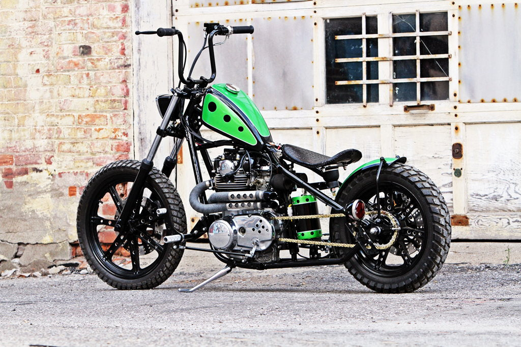 A green and black motorcycle with a TC Bros. Chrome Weld On Kick Stand for 1-1/4" Frame Tubing parked in front of a building.