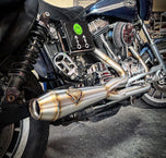 A custom SP Concepts Lane Splitter Exhaust for Harley-Davidson Dyna 1999-2005 lies on a workbench with welded joints; an American flag welding helmet is visible in the background. Stainless construction, ideal for exhaust builds.