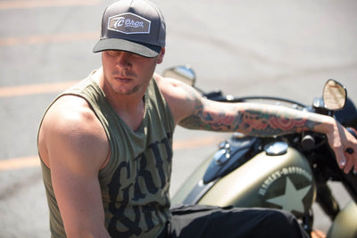 A man with tattoos sitting on a motorcycle wearing a TC Bros. Shield Snapback Hat - Charcoal/Black.
