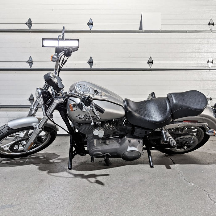 A silver motorcycle parked in front of a garage with TC Bros. Upper Shock Mount Delrin Crash Sliders 2006-2017 Harley Dyna installed.