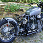 A TC Bros. vintage-style motorcycle parked in the woods with a 6-1/4" Wide Spun Steel Chopper & Bobber Fender by TC Bros Choppers.