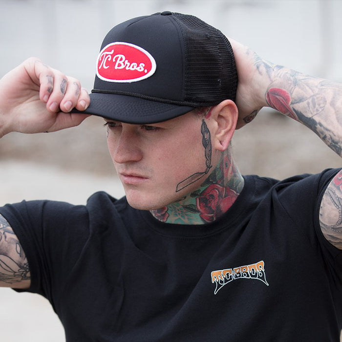 A man with tattoos wearing a black trucker hat, adorned with the TC Bros. Ol&