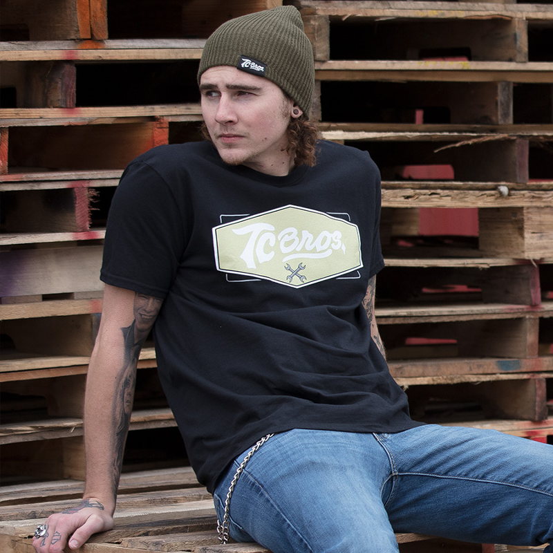 A man wearing a black t-shirt and TC Bros. Slouch Beanie - Surplus sitting on a pallet.