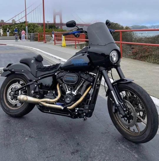 The SP Concepts Lane Splitter Exhaust for M8 Softail w/o Factory Bags—Stainless, features a 2-into-1 design, visible welds, and brand logo emblem, displayed on a workshop surface.