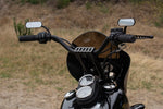 Close-up of a motorcycle’s handlebars equipped with TC Bros. Pro Series Modular Risers for Harley-Davidson, billet mirrors, and dashboard gauges; dry grass and shrubs are visible in the background.