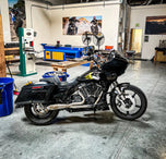 The SP Concepts Big Bore Exhaust M8 Touring FLT 2017-Present - Stainless features a polished stainless steel 2-into-1 design with visible welds and logo, resting on a gray workshop surface with an American flag-themed component in the background.