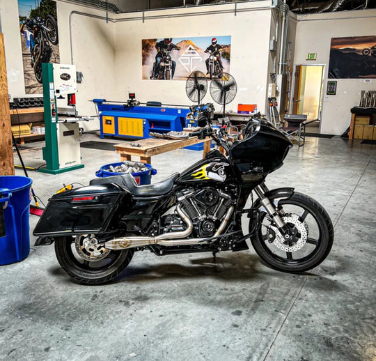 The SP Concepts Big Bore Exhaust M8 Touring FLT 2017-Present - Stainless features a polished stainless steel 2-into-1 design with visible welds and logo, resting on a gray workshop surface with an American flag-themed component in the background.