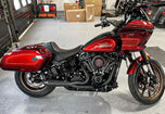 The SP Concepts Lane Splitter Exhaust for M8 Softail with Factory Bags in a stainless finish is displayed on a workbench, with visible welds and a welding helmet featuring an American flag design in the background.