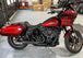 The SP Concepts Lane Splitter Exhaust for M8 Softail with Factory Bags in a stainless finish is displayed on a workbench, with visible welds and a welding helmet featuring an American flag design in the background.