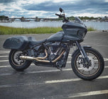 The SP Concepts Lane Splitter Exhaust for M8 Softail with Factory Bags in a stainless finish is displayed on a workbench, with visible welds and a welding helmet featuring an American flag design in the background.