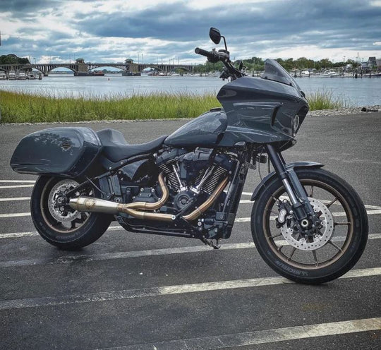 The SP Concepts Lane Splitter Exhaust for M8 Softail with Factory Bags in a stainless finish is displayed on a workbench, with visible welds and a welding helmet featuring an American flag design in the background.