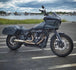 The SP Concepts Lane Splitter Exhaust for M8 Softail with Factory Bags in a stainless finish is displayed on a workbench, with visible welds and a welding helmet featuring an American flag design in the background.